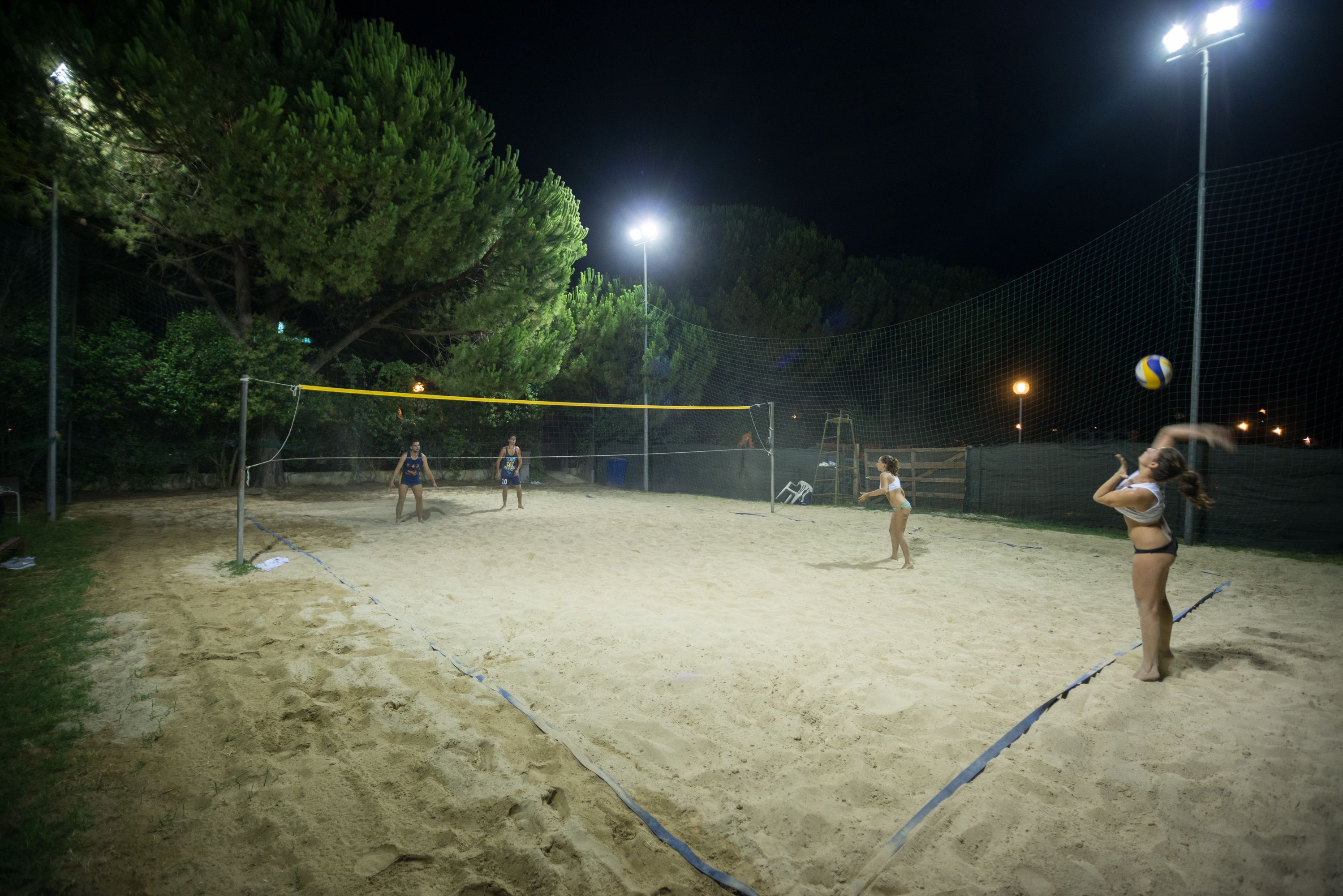 En udendørs volleyballbane om natten med net, sand og træer i baggrunden.