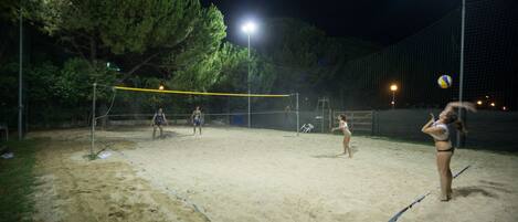 En udendørs volleyballbane om natten med net, sand og træer i baggrunden.