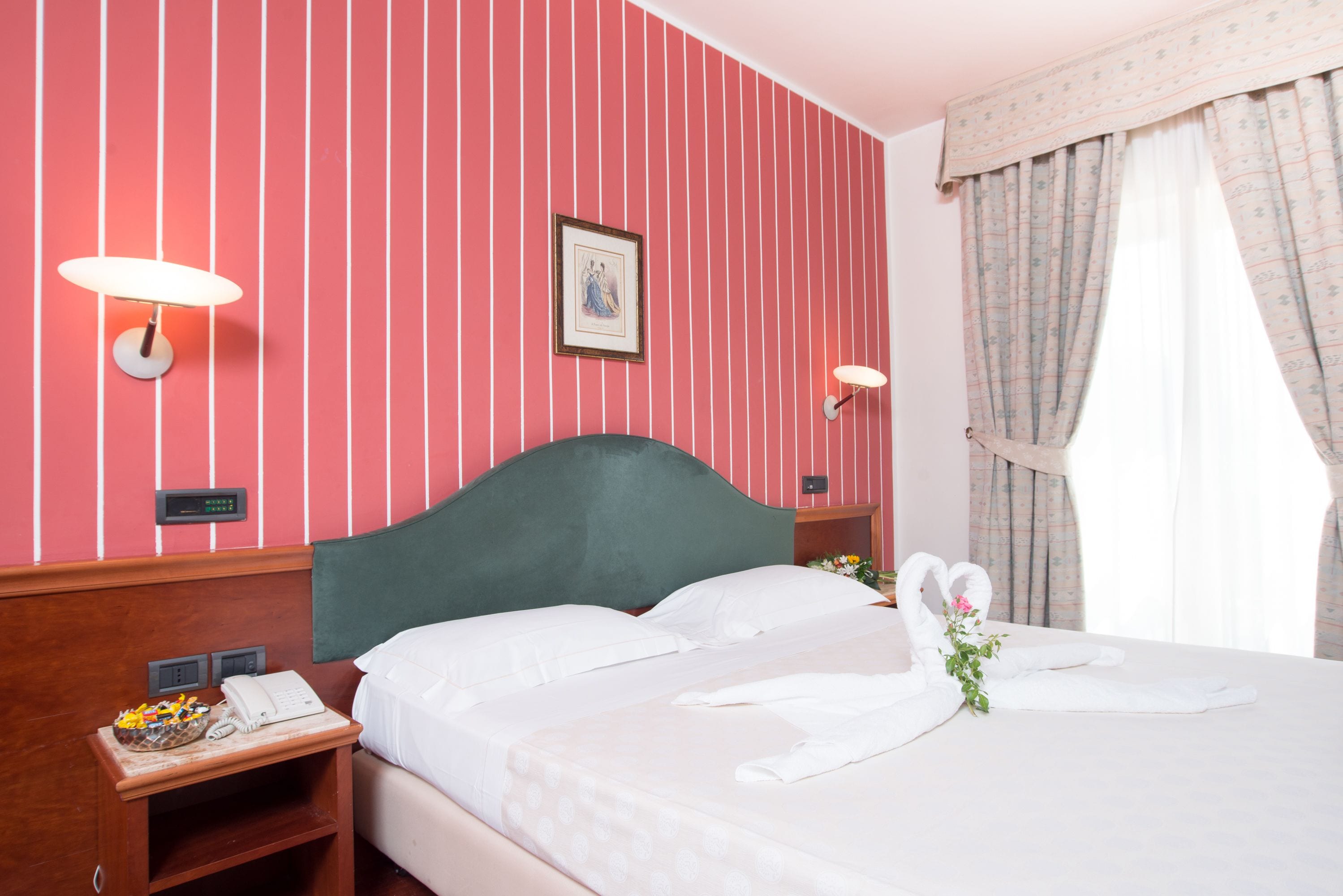 Une chambre d’hôtel avec un mur à rayures roses, une tête de lit verte, une table de chevet avec un téléphone et un bouquet de fleurs sur le lit.