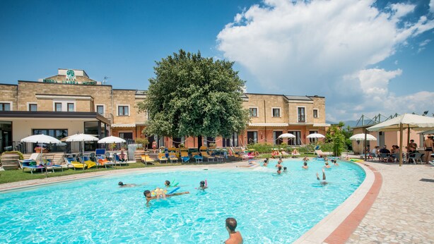 Una piscina con sillas de descanso y sombrillas, rodeada por un edificio con un árbol grande.