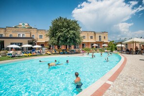 Una piscina con sillas de descanso y sombrillas, rodeada por un edificio con un árbol grande.