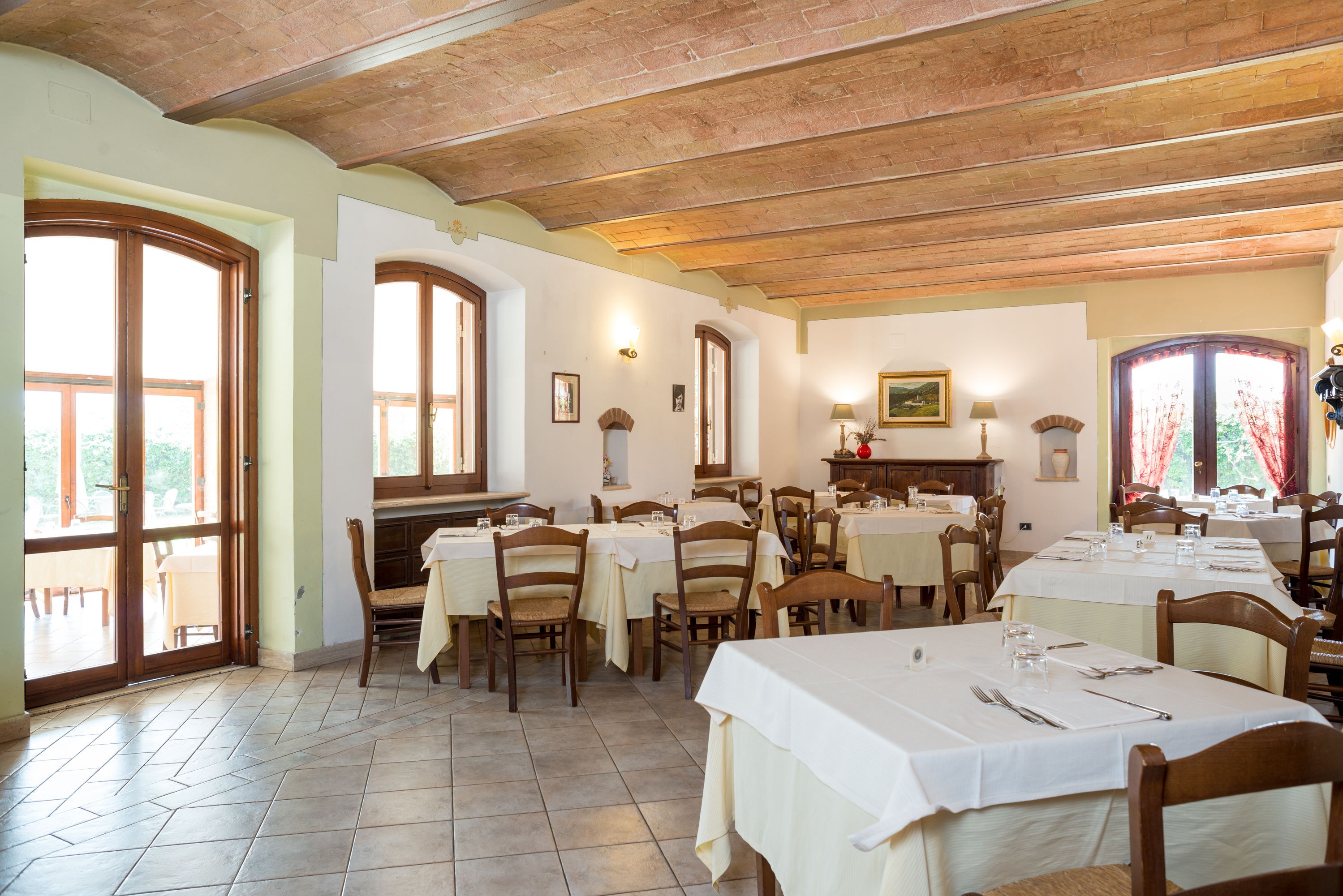 A dining area with tables set for service, wooden chairs, and large windows allowing natural light.