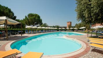 Area piscina con lettini e ombrelloni, circondata da alberi e un edificio sullo sfondo.