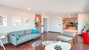 Un salón moderno con un sofá gris, una mesita redonda pequeña y un sillón gris. Hay una estantería de madera con objetos decorativos y una planta en maceta. El suelo es de madera y las paredes son blancas.