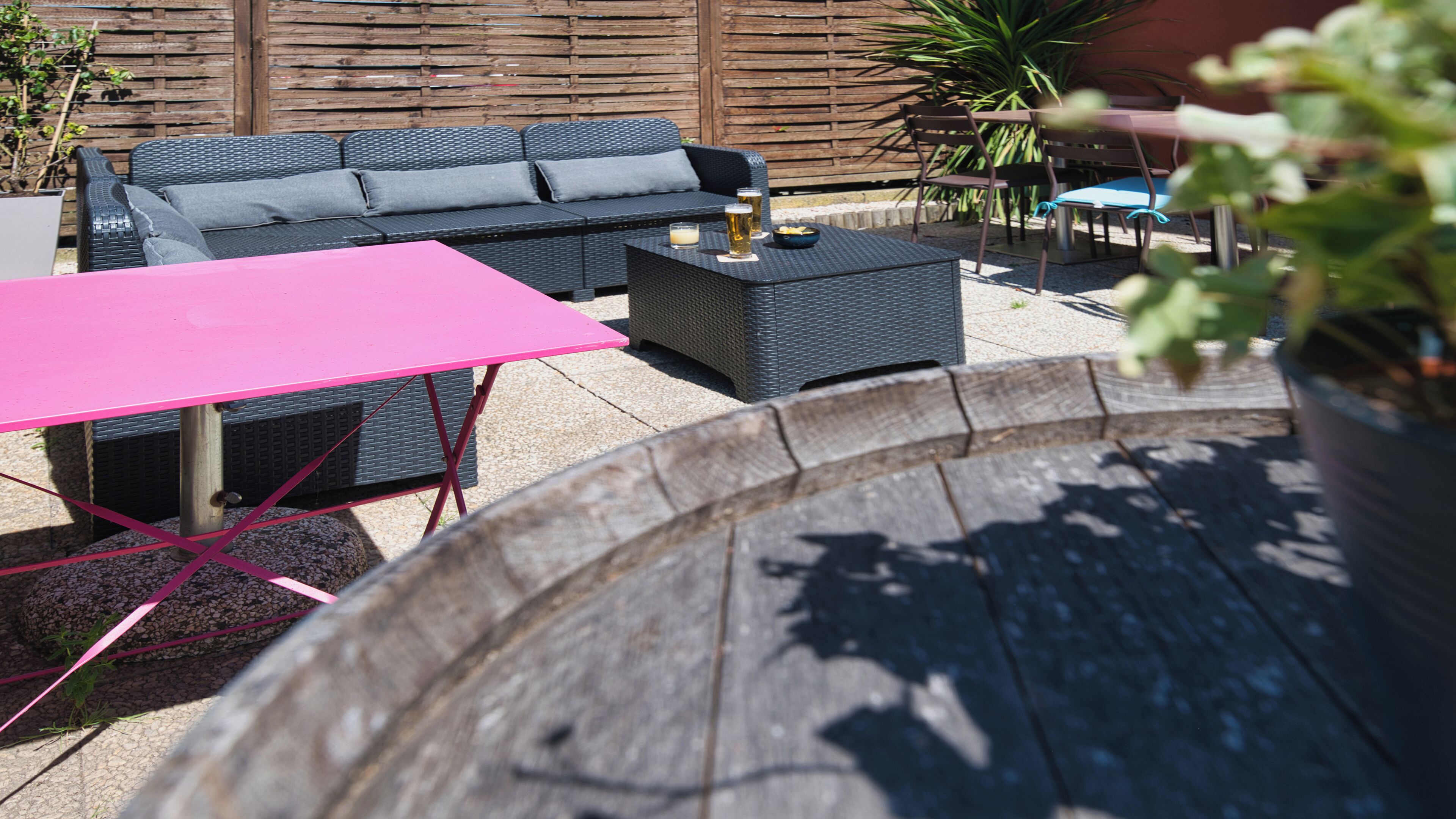 Un espace patio aménagé avec un canapé en osier noir, une table rose et un bac à fleurs rond en bois.