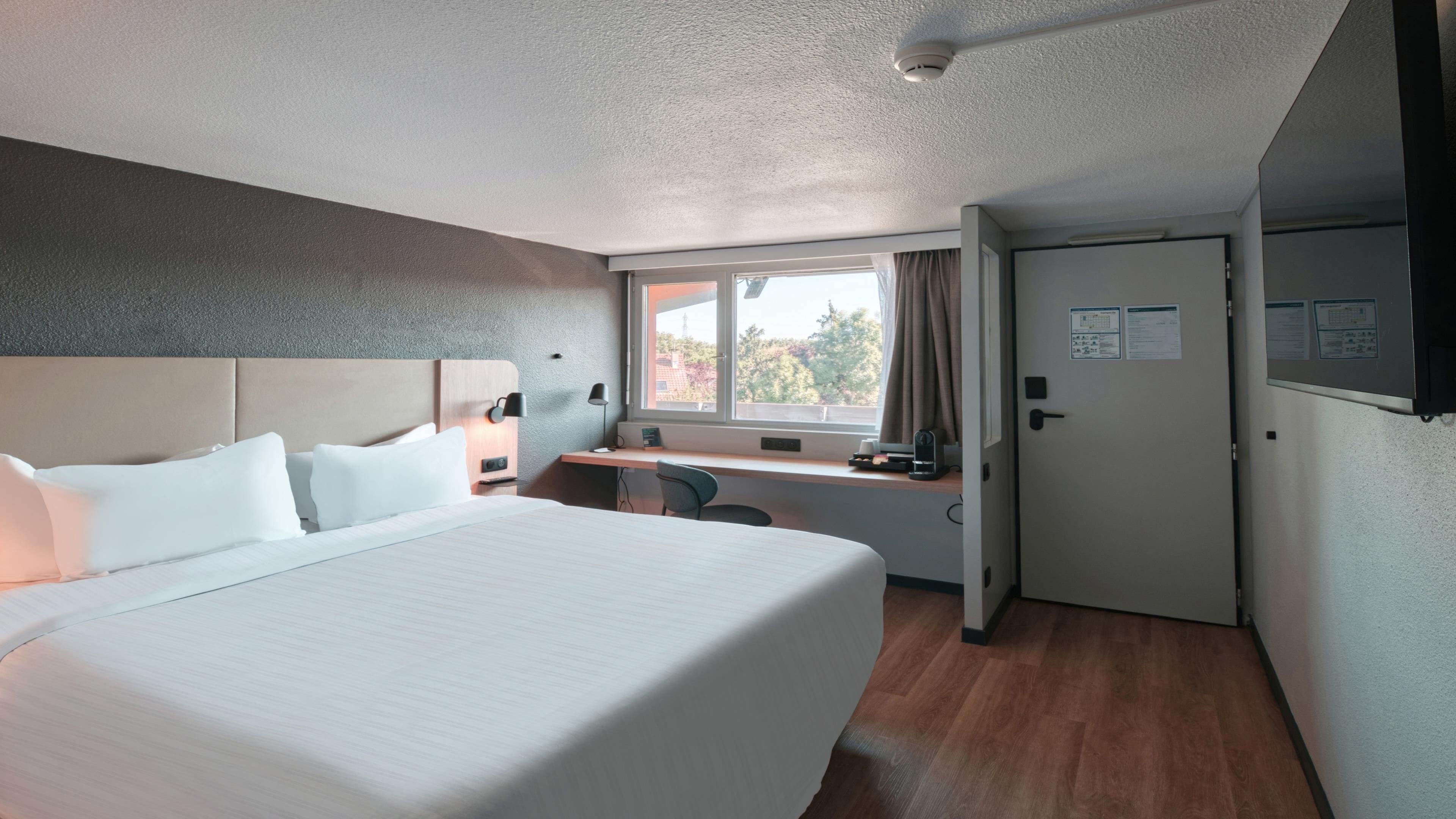 A hotel room with a bed, desk, and window.