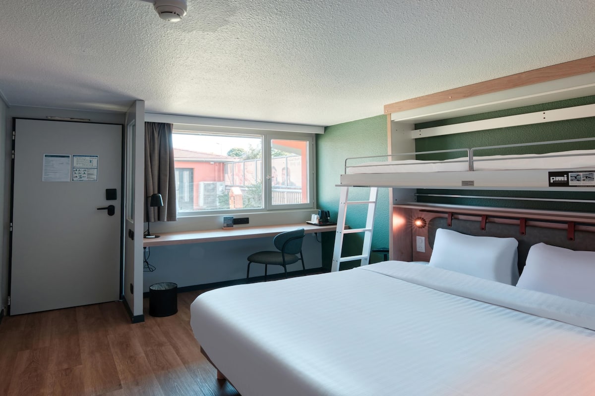 Une chambre d’hôtel compacte comprenant un lit superposé, un bureau avec une chaise, une fenêtre avec des rideaux et une porte sur laquelle est affiché un avis.