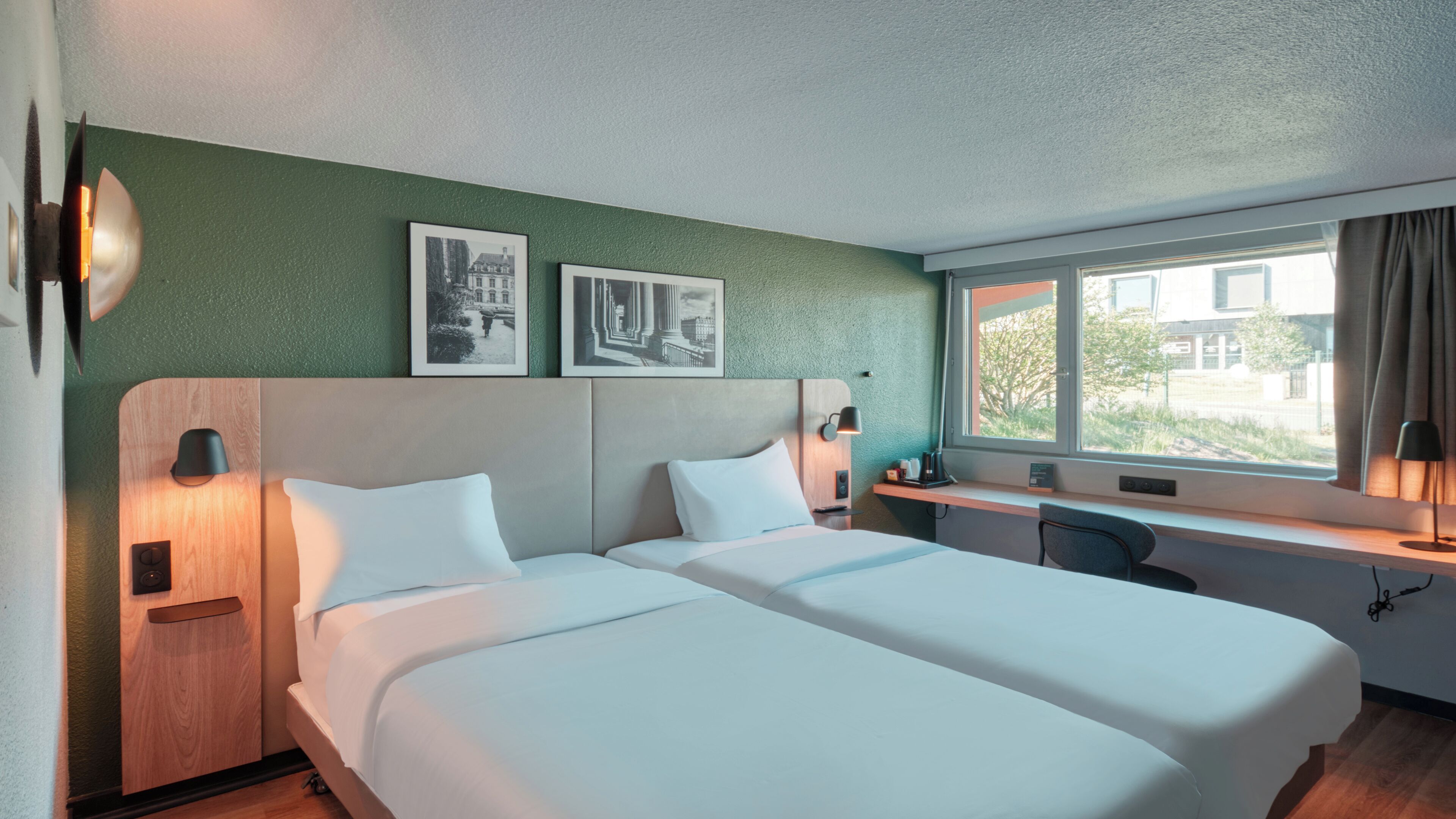 Een hotelkamer met twee bedden, een bureau, een stoel en een raam met uitzicht op gebouwen.
