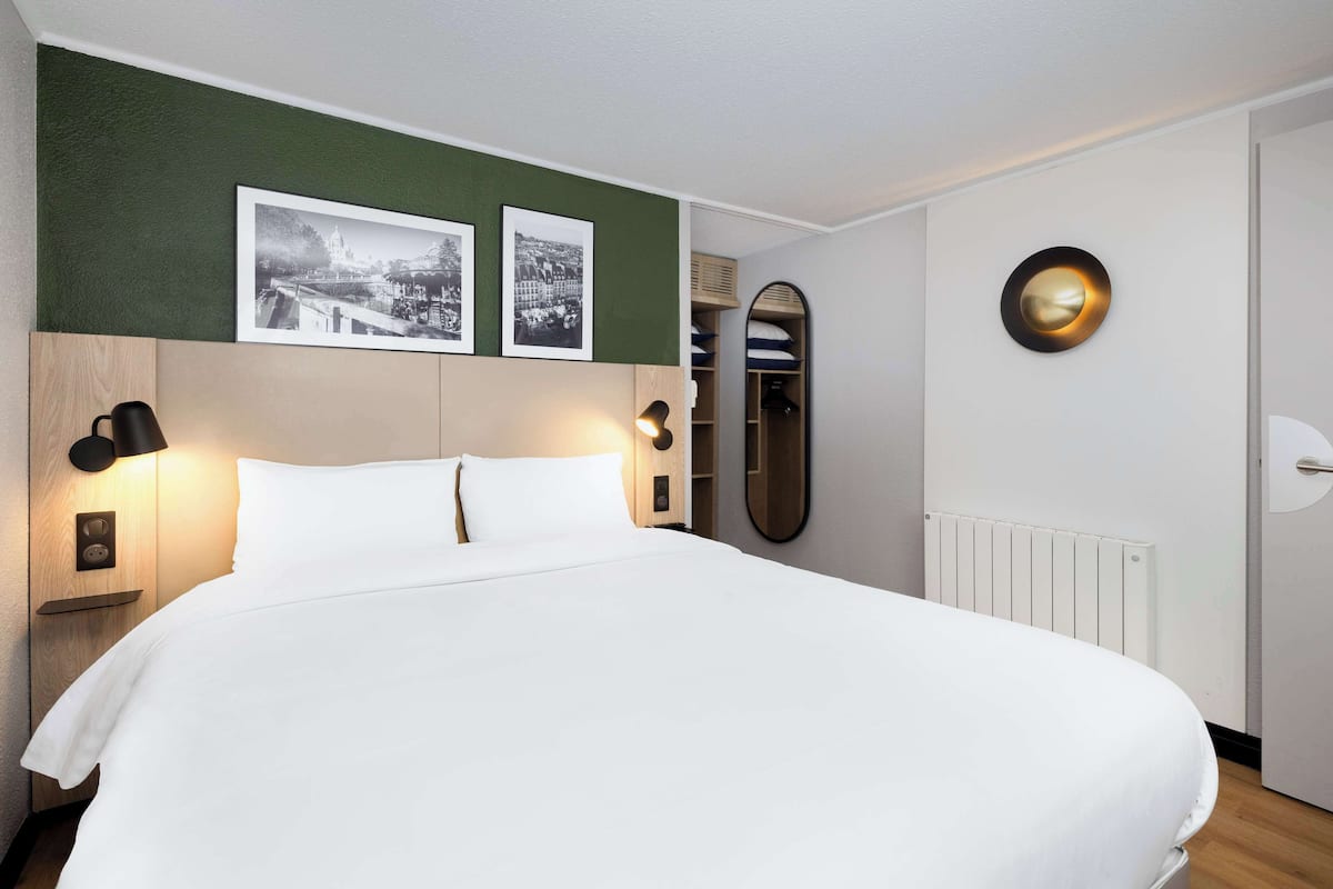 Un lit bien fait, avec du linge de lit blanc, une tête de lit en bois, deux lampes fixées au mur et deux photographies en noir et blanc encadrées au-dessus du lit.