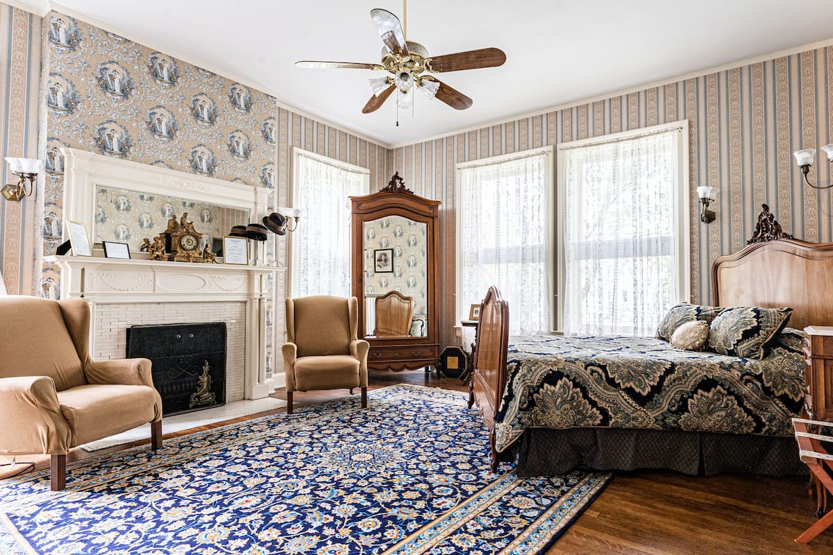A room with a fireplace, a large bed, a wooden chair, a mirror, and a patterned rug.