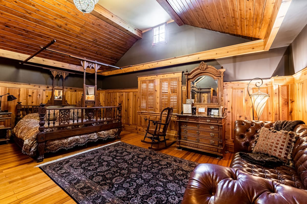 A room with a wooden ceiling, a large bed, a leather sofa, a rocking chair, and a dresser with a mirror.