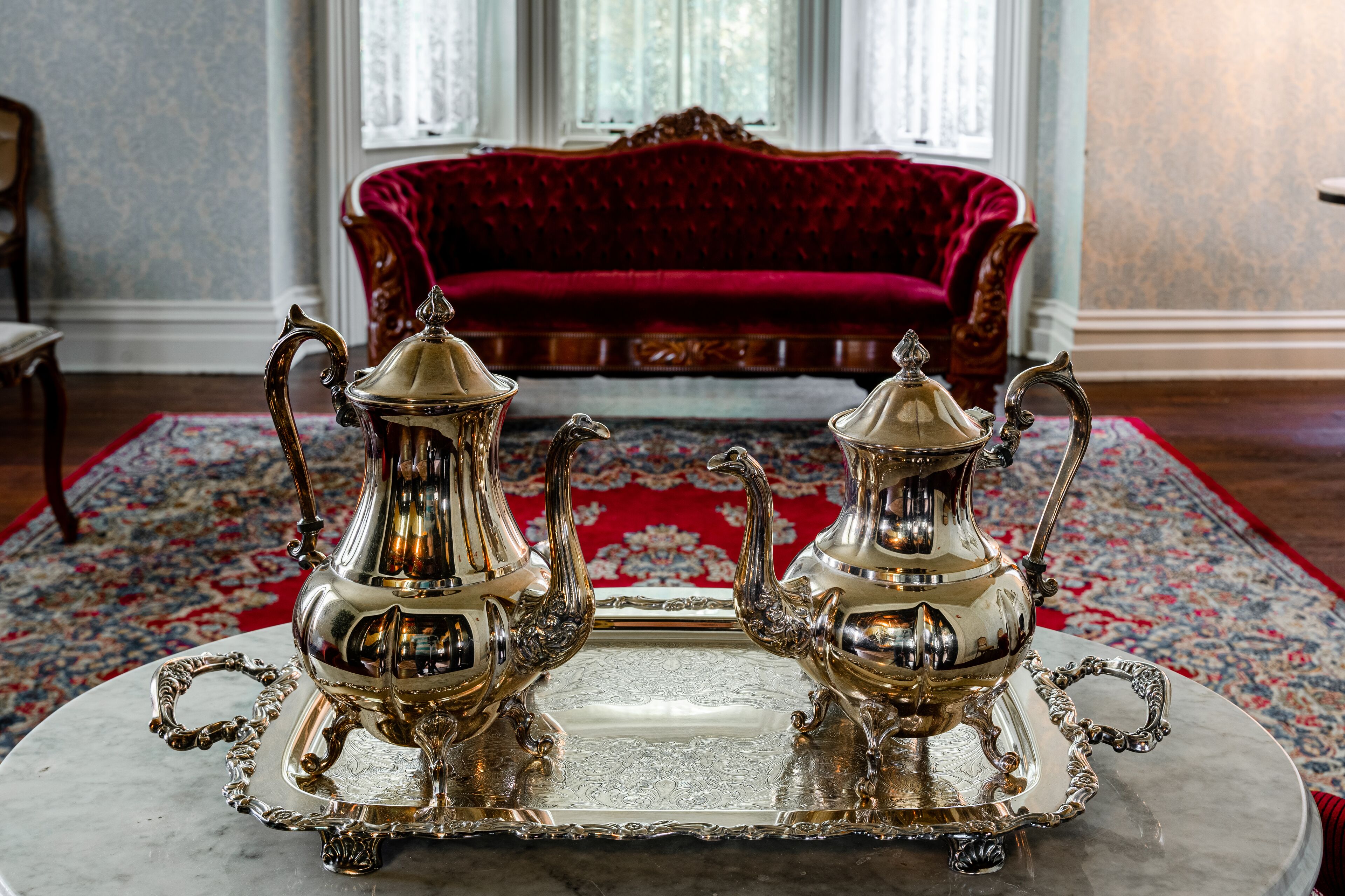 Ein Zimmer mit einem roten Samtsofa, einem gemusterten Teppich und zwei verzierten Silber-Teekannen auf einem Marmortisch.