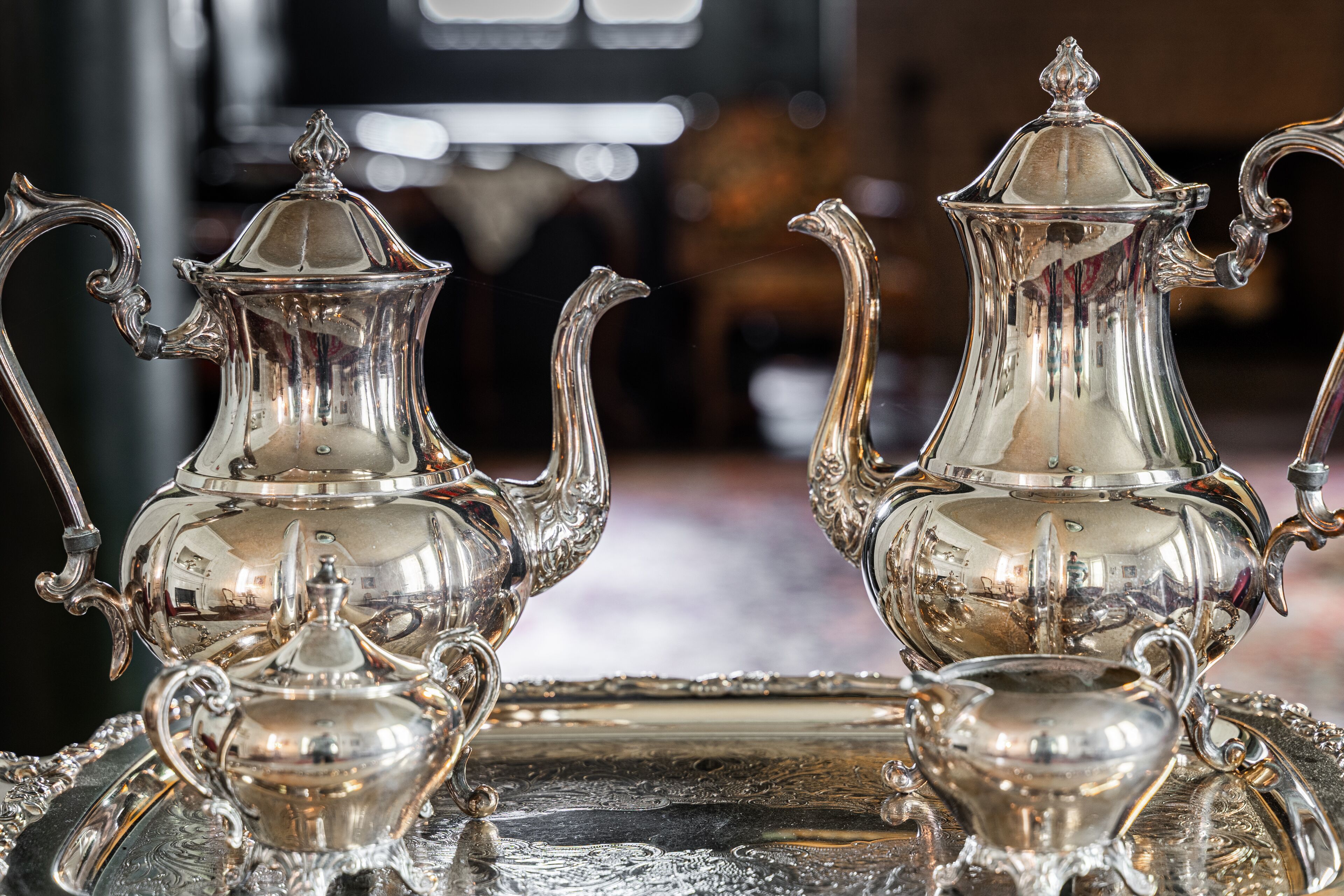 Deux théières en argent richement décorées, dotées d’anses et de couvercles, sont posées sur un plateau réfléchissant.