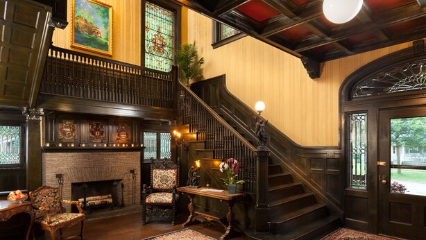 A grand staircase with a chandelier, a fireplace, and ornate wall decorations in a spacious room with a large rug. - Vrooman Mansion B&B (Bloomington)