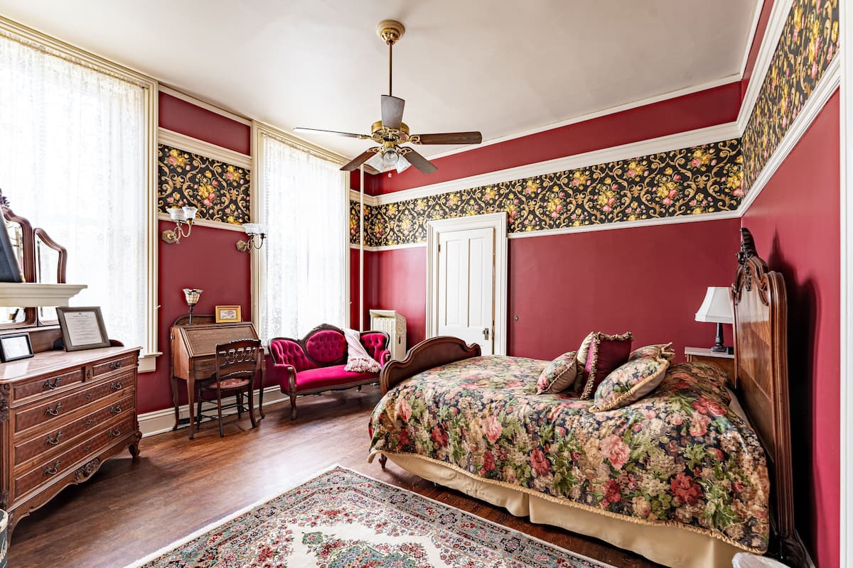 A bedroom with a wooden dresser, a desk, a chair, a bed with floral bedding, a ceiling fan, and patterned wallpaper.