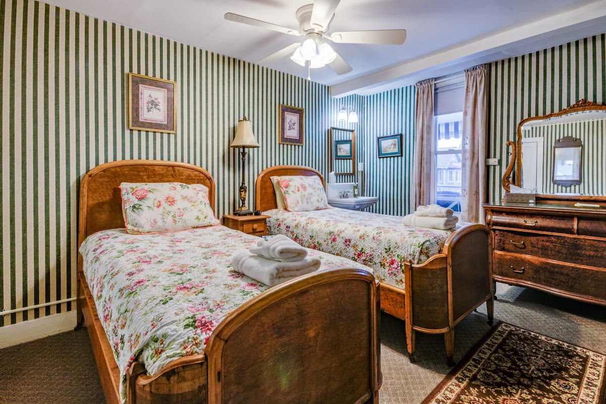 A hotel room with two wooden beds, striped wallpaper, a ceiling fan, a dresser, and a mirror.