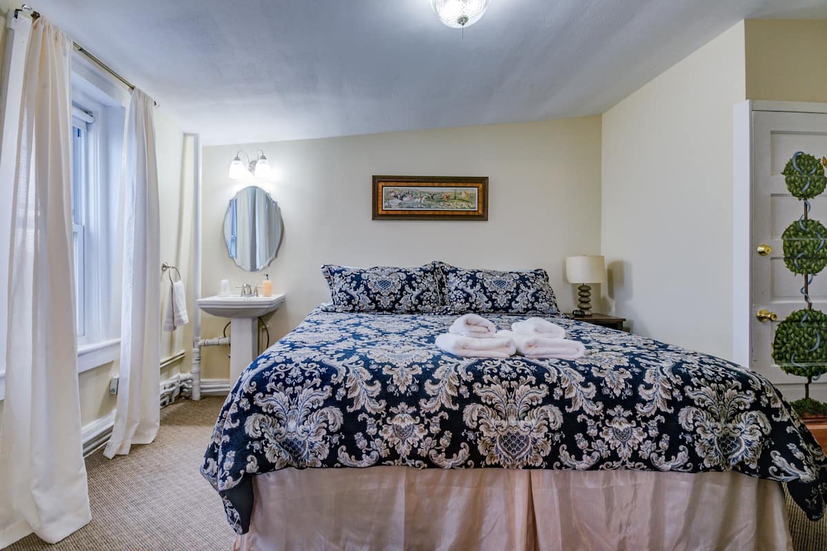 A neatly arranged bedroom with a large bed, a bedside table with a lamp, a mirror, and a framed picture on the wall.