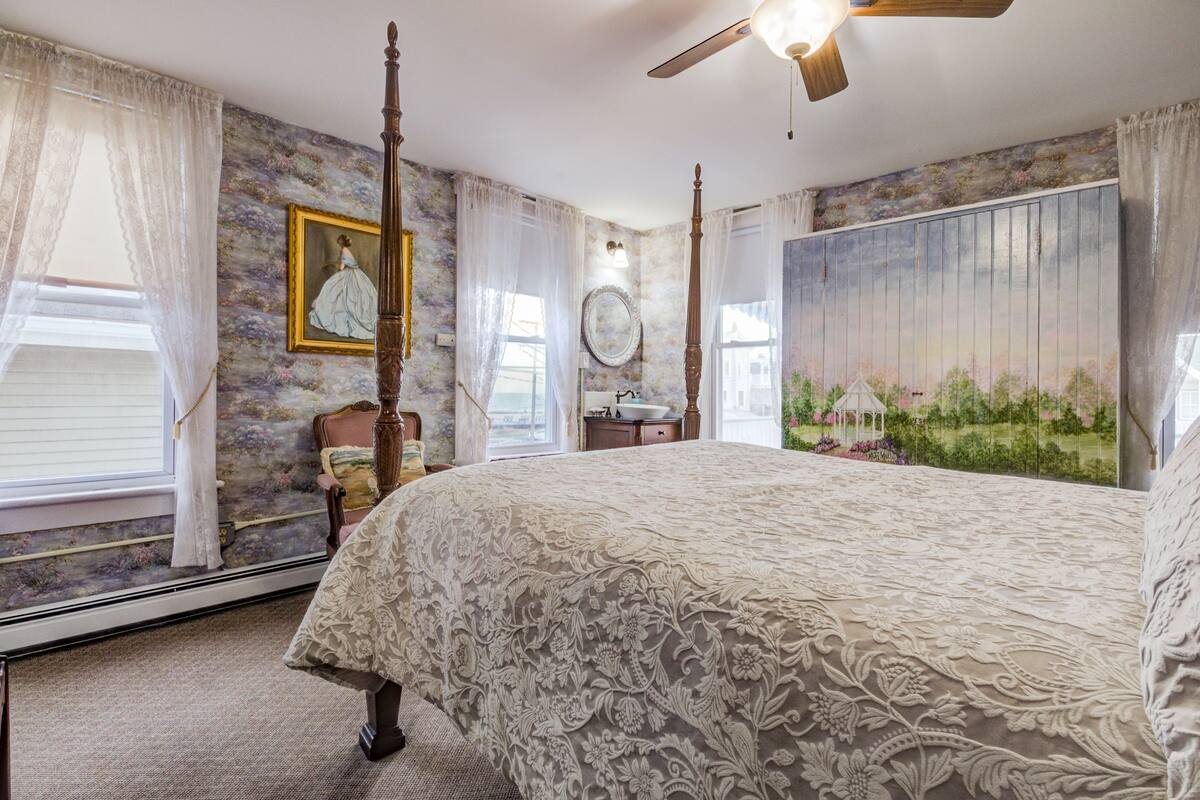 A bedroom with a four-poster bed, a ceiling fan, and a large window with curtains.