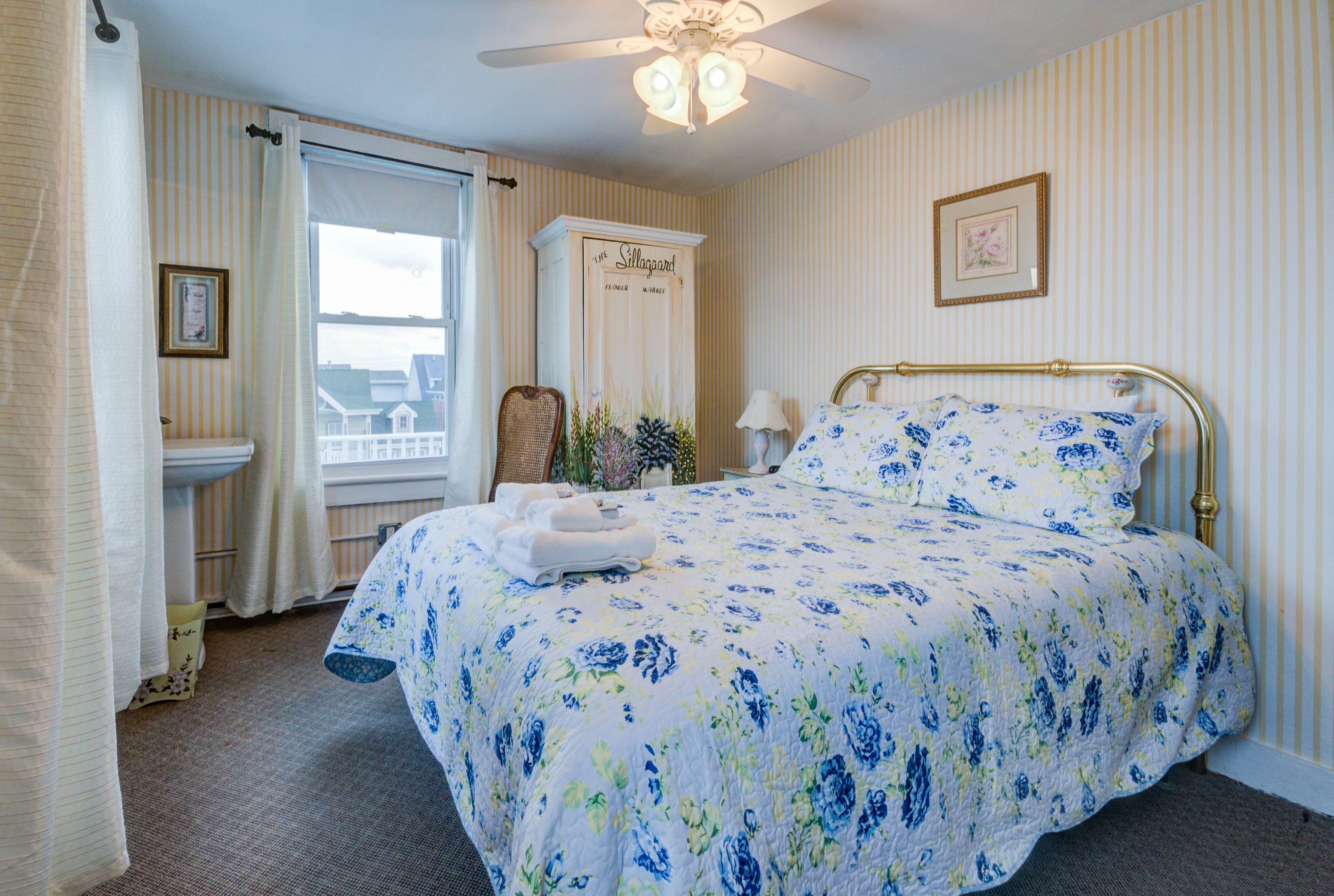 Una habitación de hotel con una cama grande, una ventana con cortinas, un ventilador de techo, una mesita de noche con una lámpara y un cuadro en la pared.