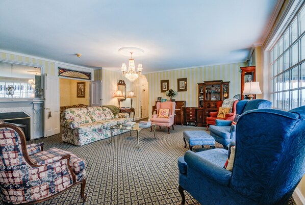 A spacious living room with a patterned carpet, a chandelier, and a variety of seating options including a blue armchair, a floral sofa, and a pink armchair.