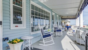 A covered porch with white rocking chairs and tables, a potted plant, and a view of the ocean. - The Lillagaard B&B (Ocean Grove)