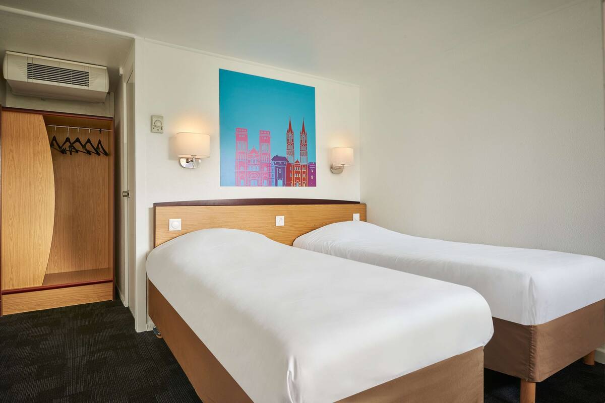 Habitación de hotel con dos camas individuales, un armario de madera, perchero y un cuadro de un paisaje urbano en la pared.