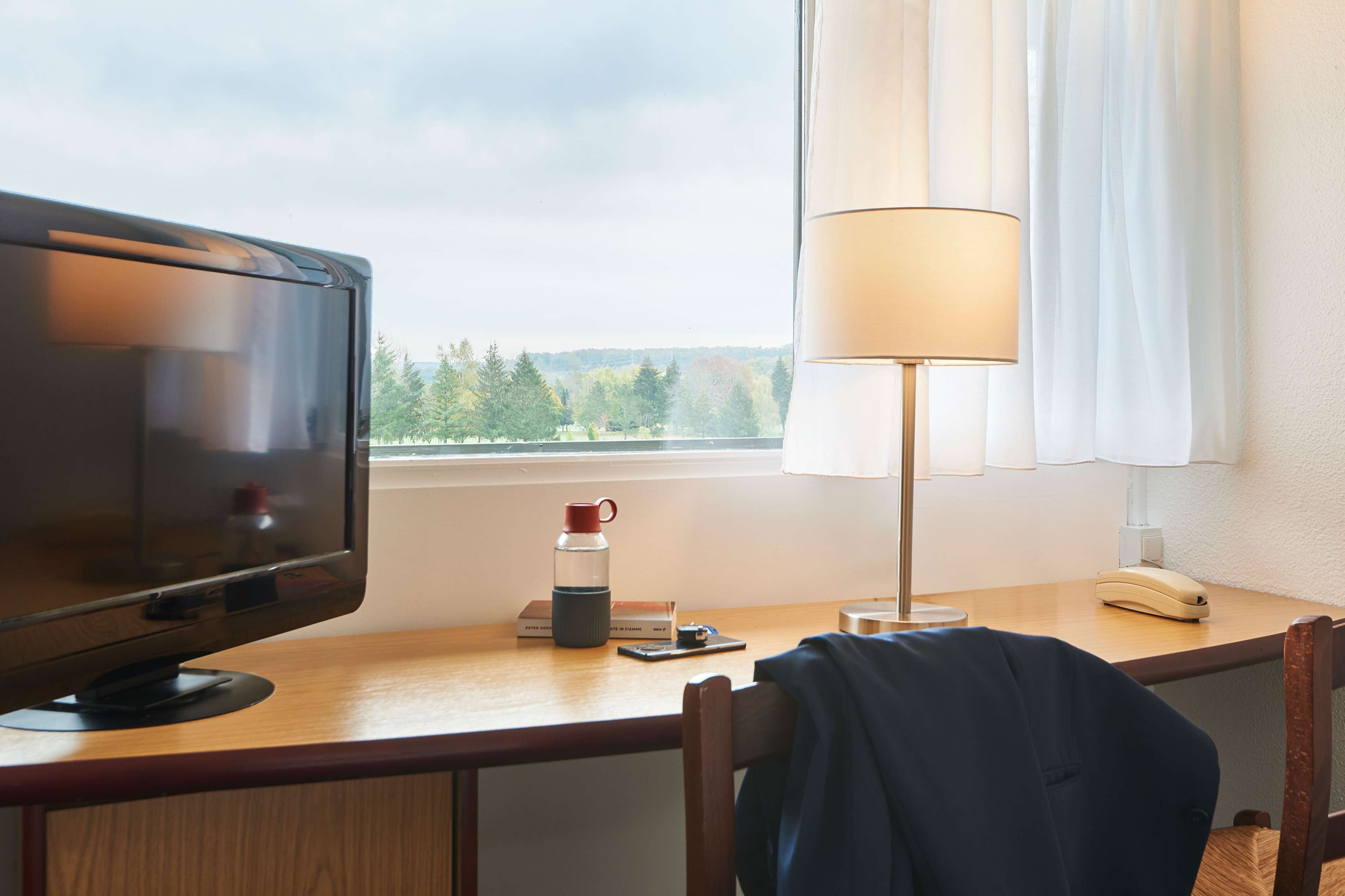 Ett hotellrum med ett skrivbord, en tv, en lampa, en telefon och ett fönster med utsikt över träd.