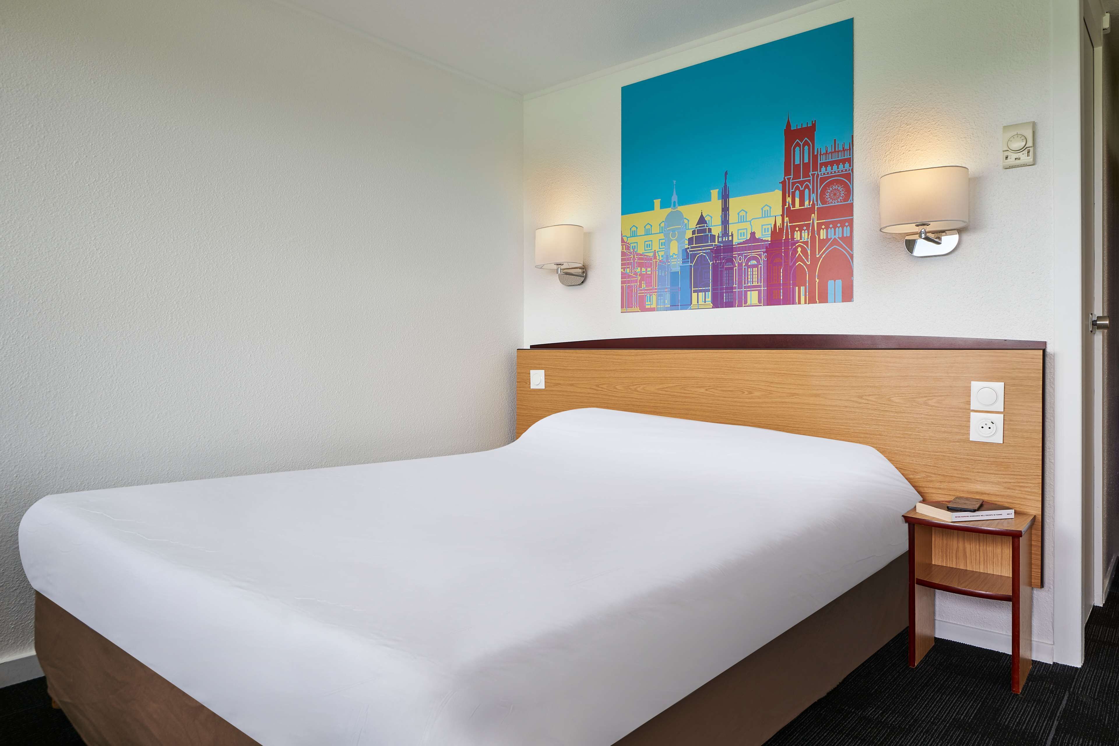 Een hotelkamer met een groot bed, een houten hoofdbord, twee wandlampen en een kleurrijk stadsgezicht schilderij.