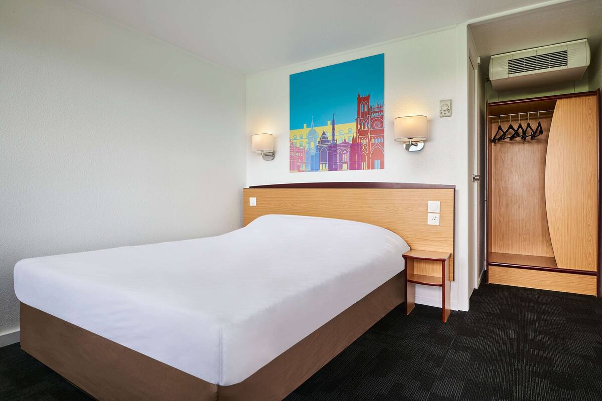 Habitación de hotel con una cama grande, una mesita de noche pequeña, un armario para abrigos y un cuadro de la ciudad en la pared.