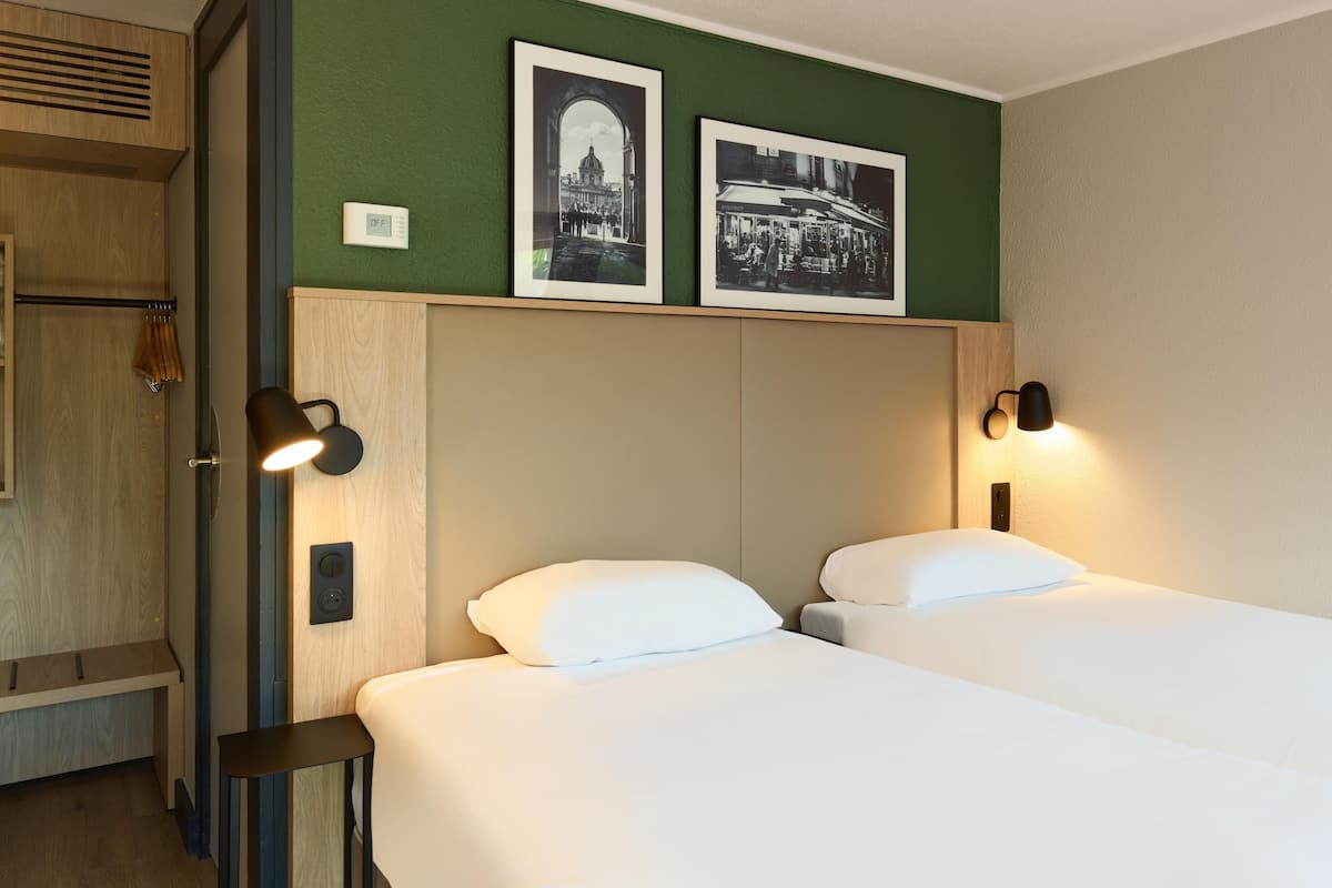 A hotel room with two beds, a wooden headboard, two wall-mounted lamps, and two framed pictures on the wall.
