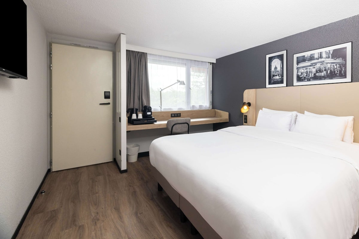 A hotel room with a bed, a desk, a chair, a window with curtains, and two framed pictures on the wall.