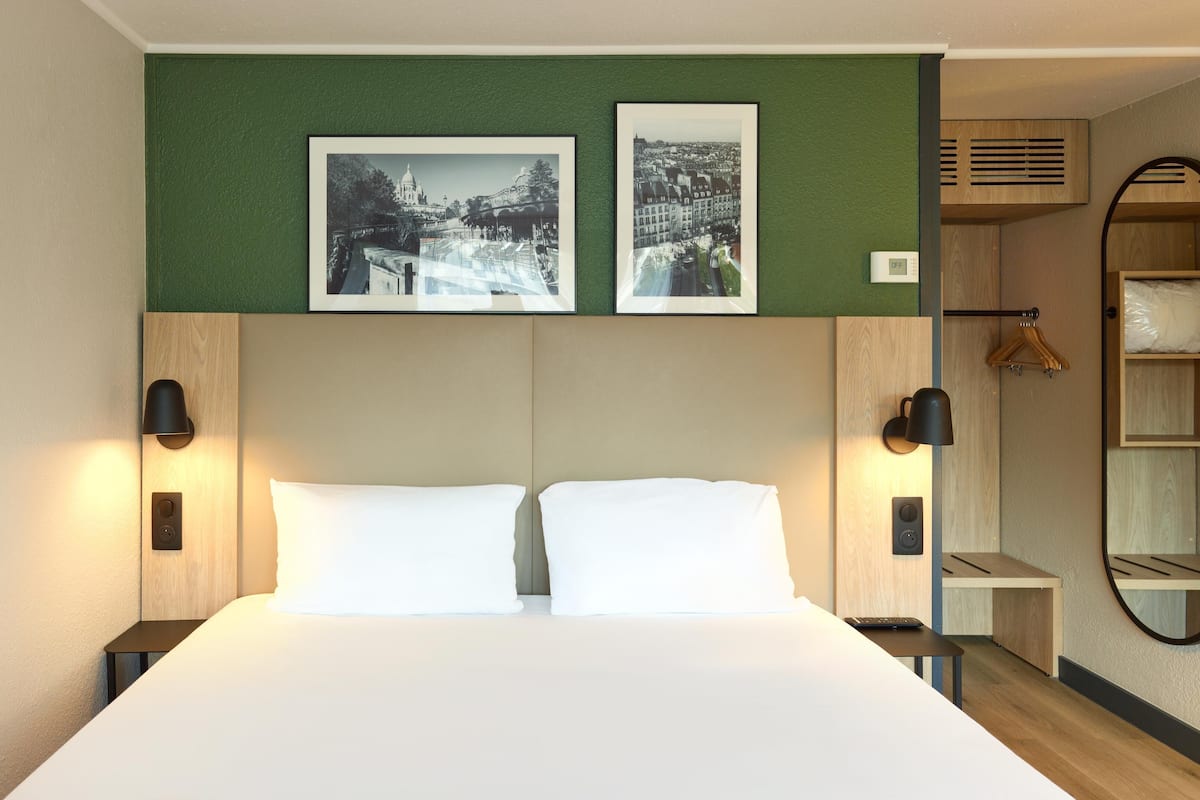 A hotel room with a large bed, two wall-mounted lamps, a side table, a mirror, and two framed pictures above the bed.