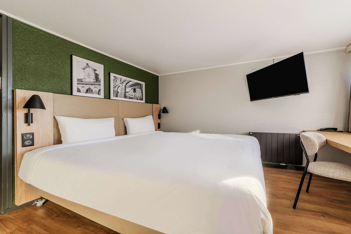 Ein Hotelzimmer mit einem großen Bett, einem Schreibtisch mit Stuhl, einem flachbildfernseher an der Wand und zwei gerahmten Bildern über dem Bett.