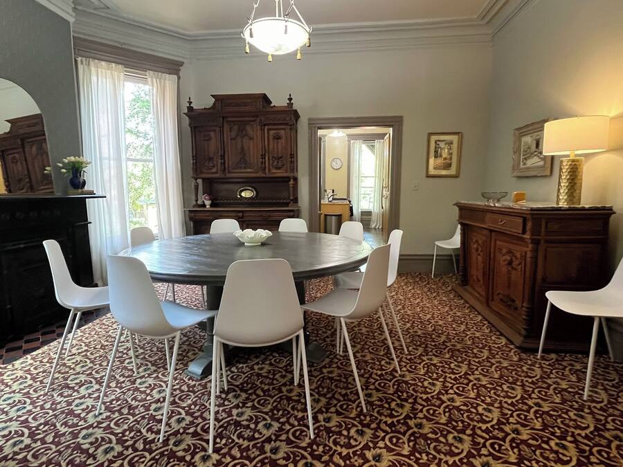 Un comedor con una mesa redonda, sillas blancas, una alfombra estampada, un aparador de madera y una araña.