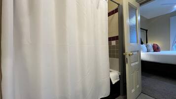 A hotel room with a bed, a bathroom with a shower curtain, and a door leading to another room.