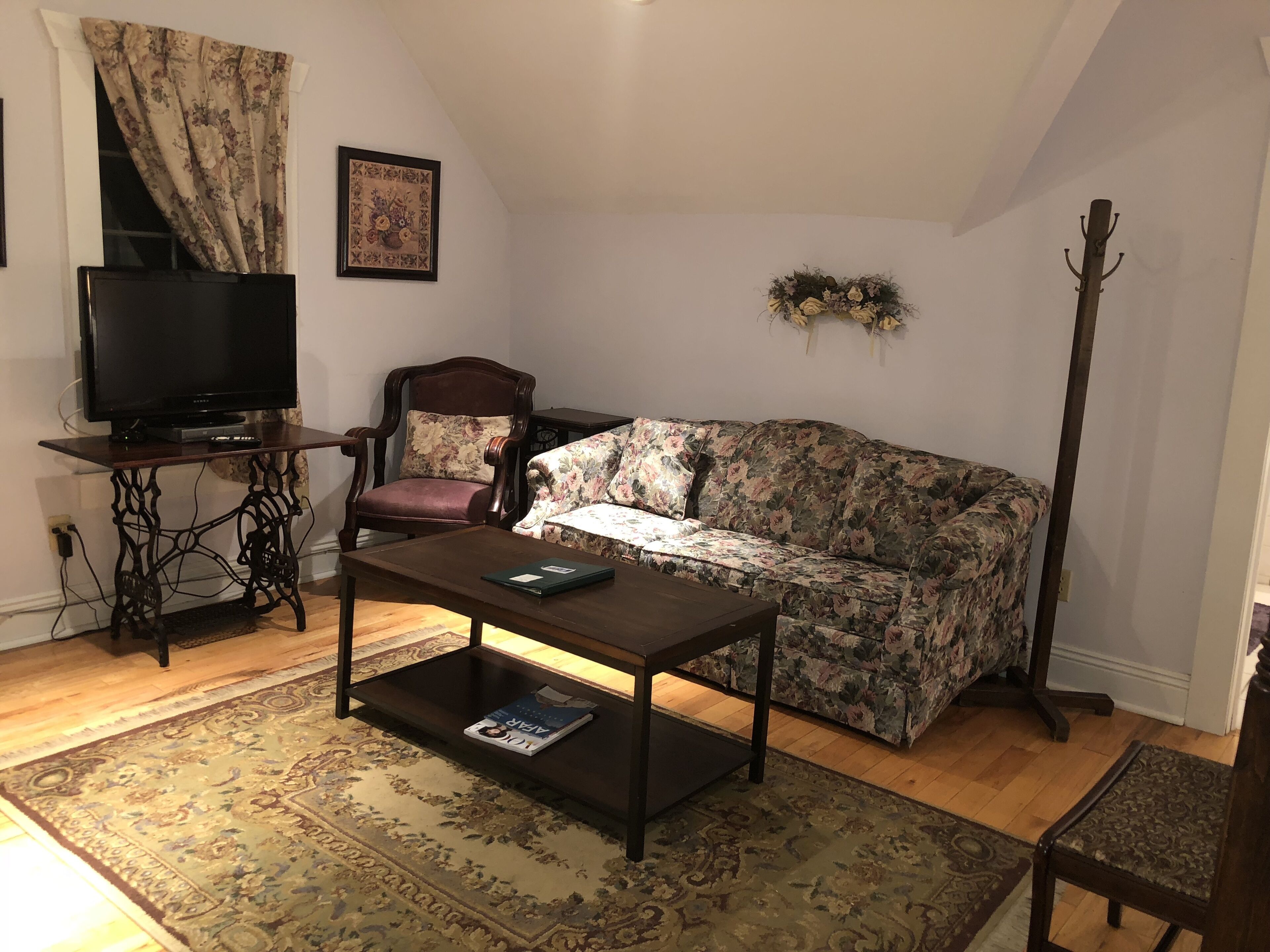 Un salon avec une table basse en bois foncé, un canapé à motifs floraux, un fauteuil en bois, un téléviseur et un porte-manteau.