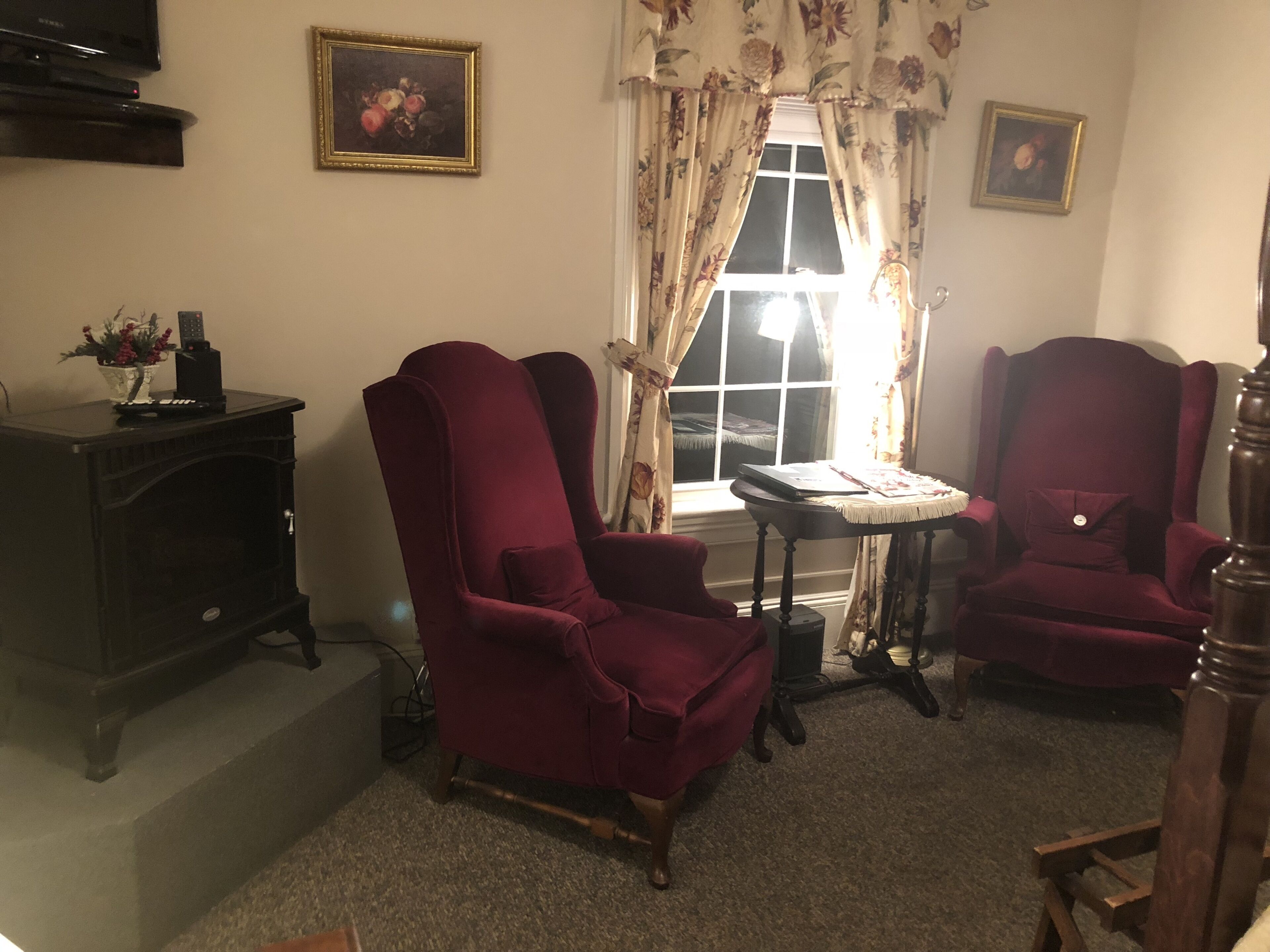 Una sala de estar con dos sillones rojos, una mesita, un aparador de madera oscura y una ventana con cortinas florales.