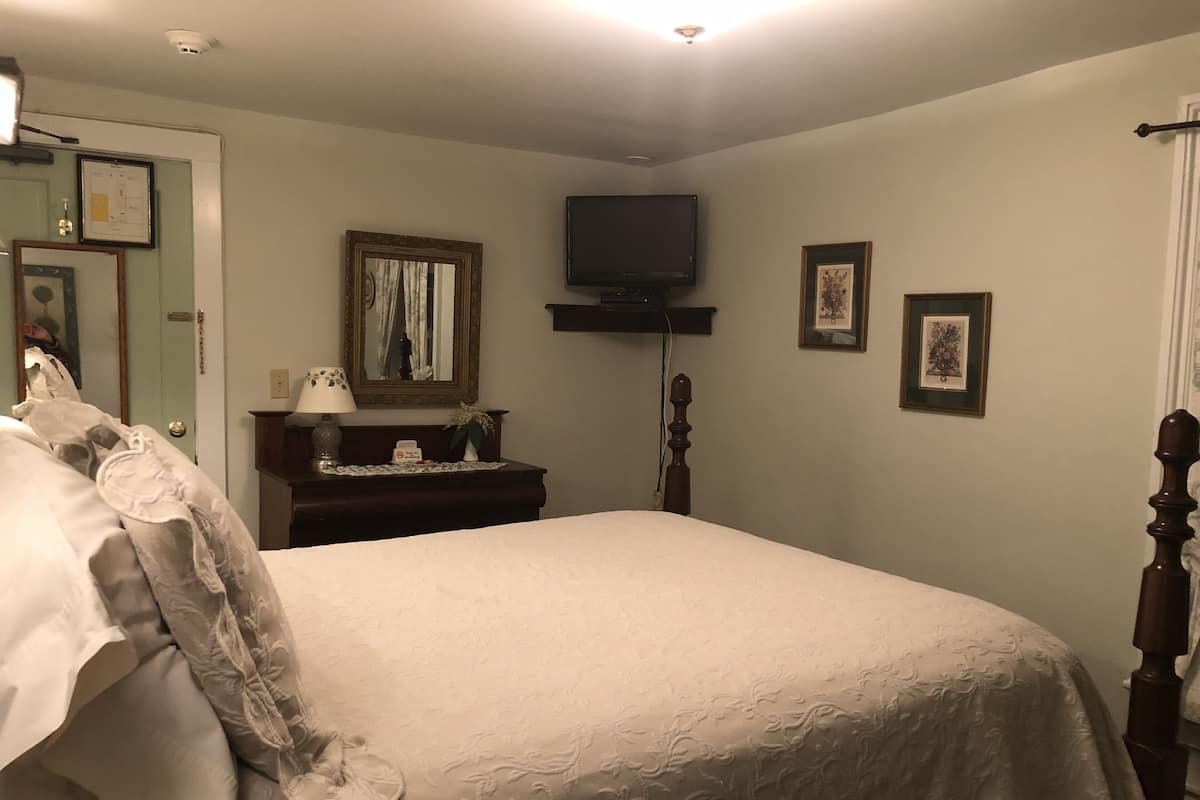 A neatly made bed with a white bedspread, a bedside table with a lamp, a mirror, and a television mounted on the wall.