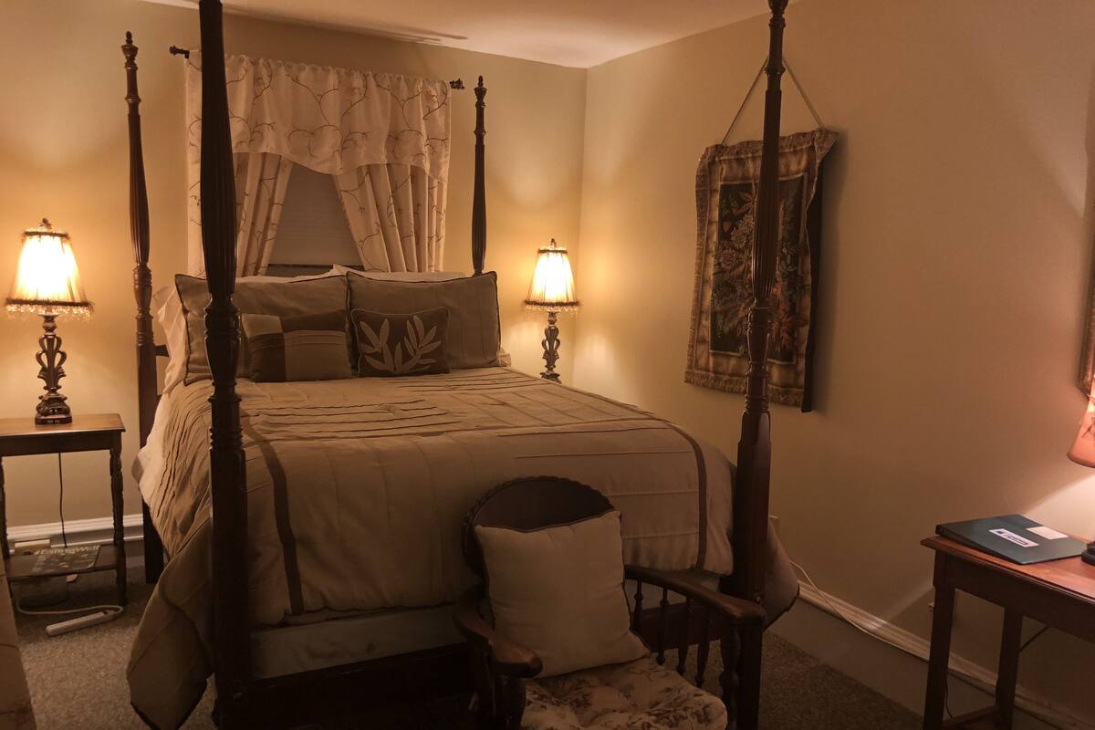 A four-poster bed with a canopy, two bedside lamps, a chair with a cushion, and a small table with a book.