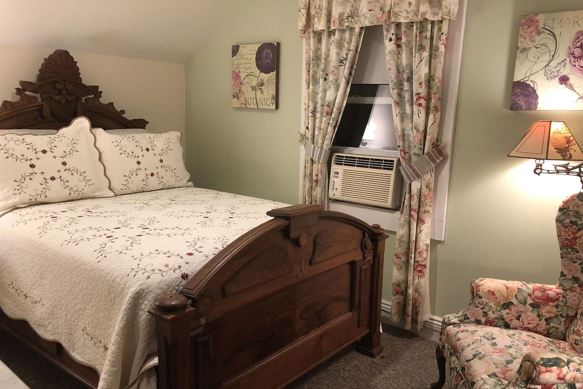 A bedroom with a wooden bed, floral curtains, a radiator, and a floral armchair.