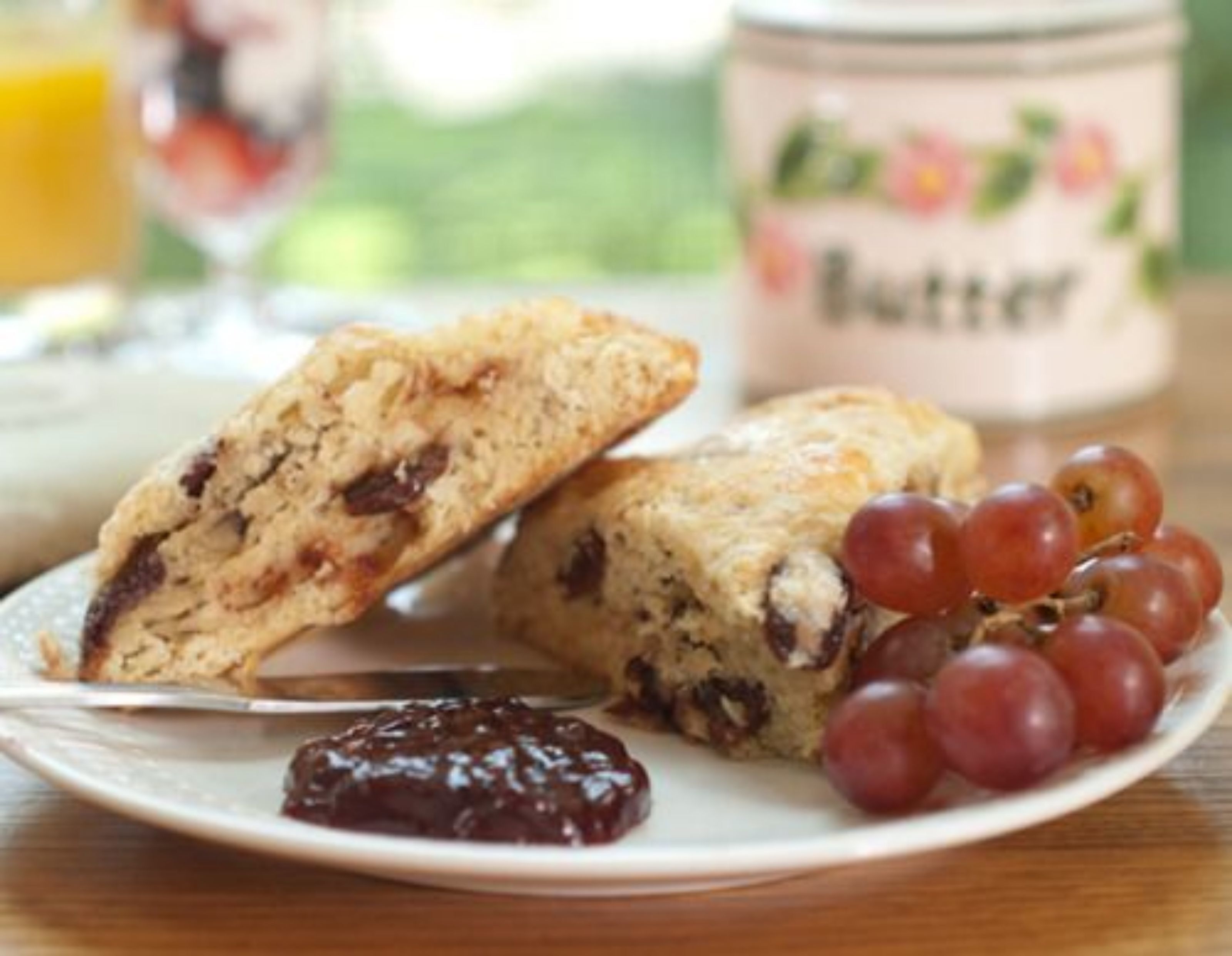 A plate with two scones, a jar of jam, and a bunch of grapes.