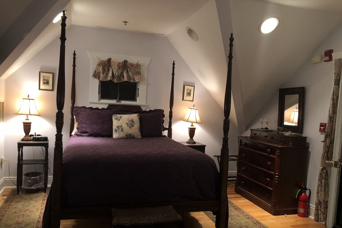 A four-poster bed with a purple bedspread, two bedside lamps, a wooden dresser, a fire extinguisher, and a window with a curtain.