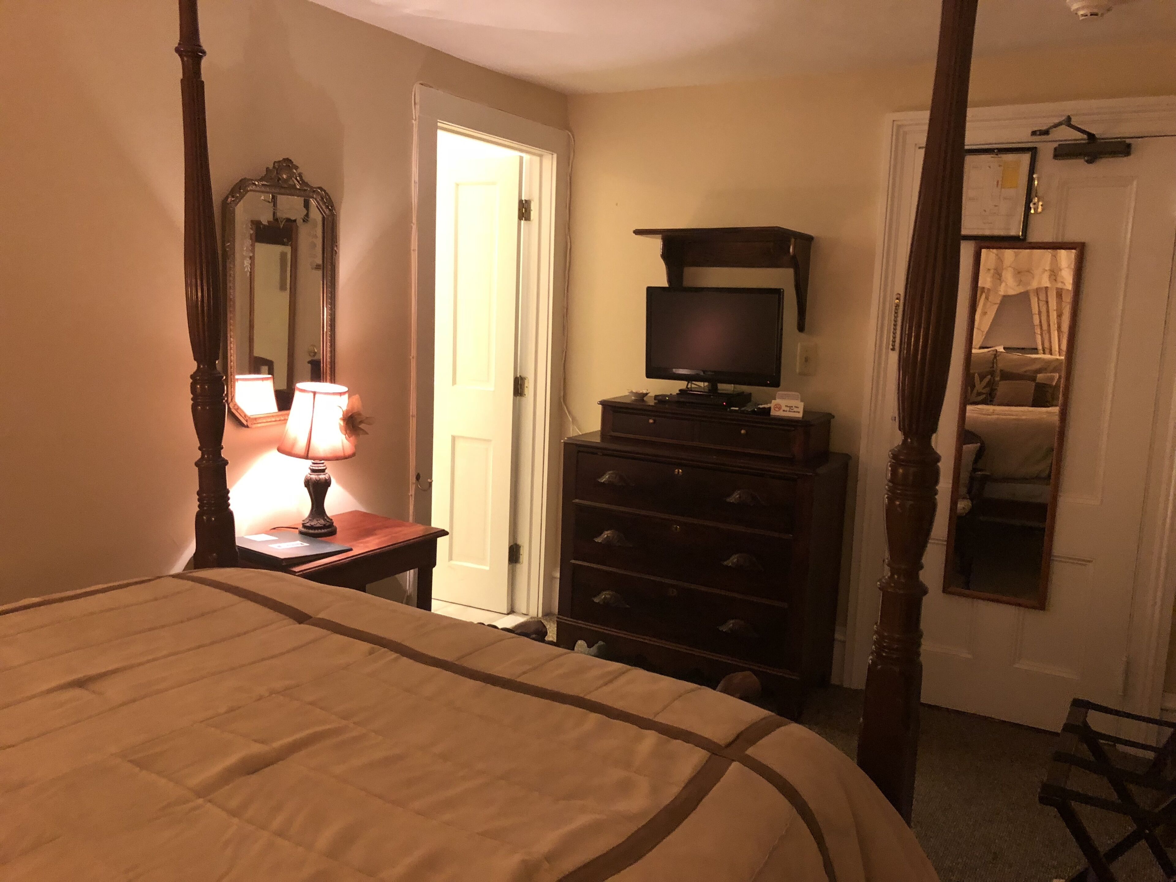 Ein Schlafzimmer mit Himmelbett, Kommode, einem Fernseher darauf und einem Spiegel an der Wand.