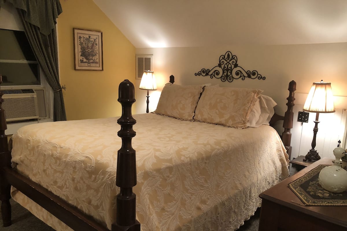 A bedroom with a large bed, two bedside lamps, a decorative wall headboard, a framed picture, and a window with curtains.