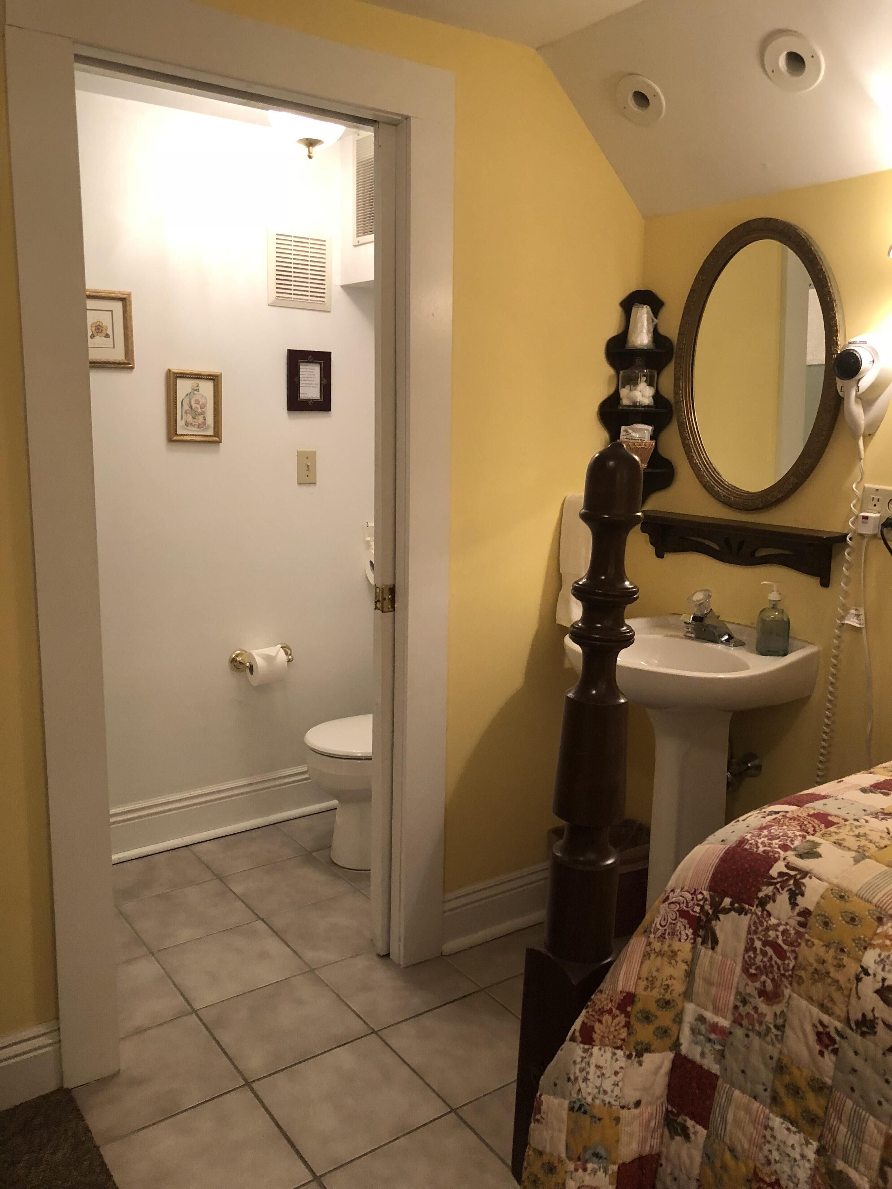Un baño con inodoro, lavamanos y espejo. Hay una puerta que da a otra habitación.