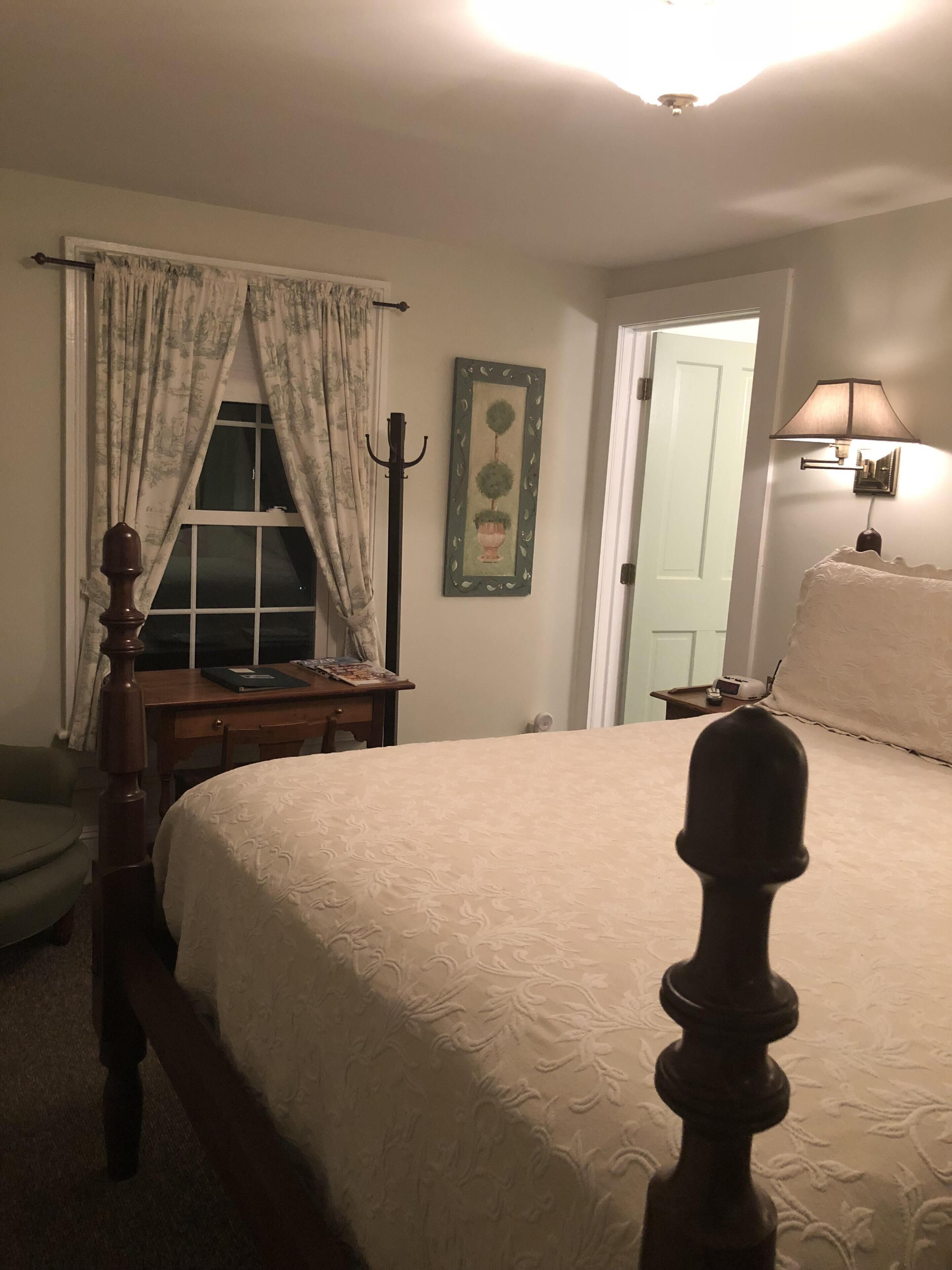 Un dormitorio con cama con dosel, una mesita de noche con lámpara, una ventana con cortinas y una puerta.