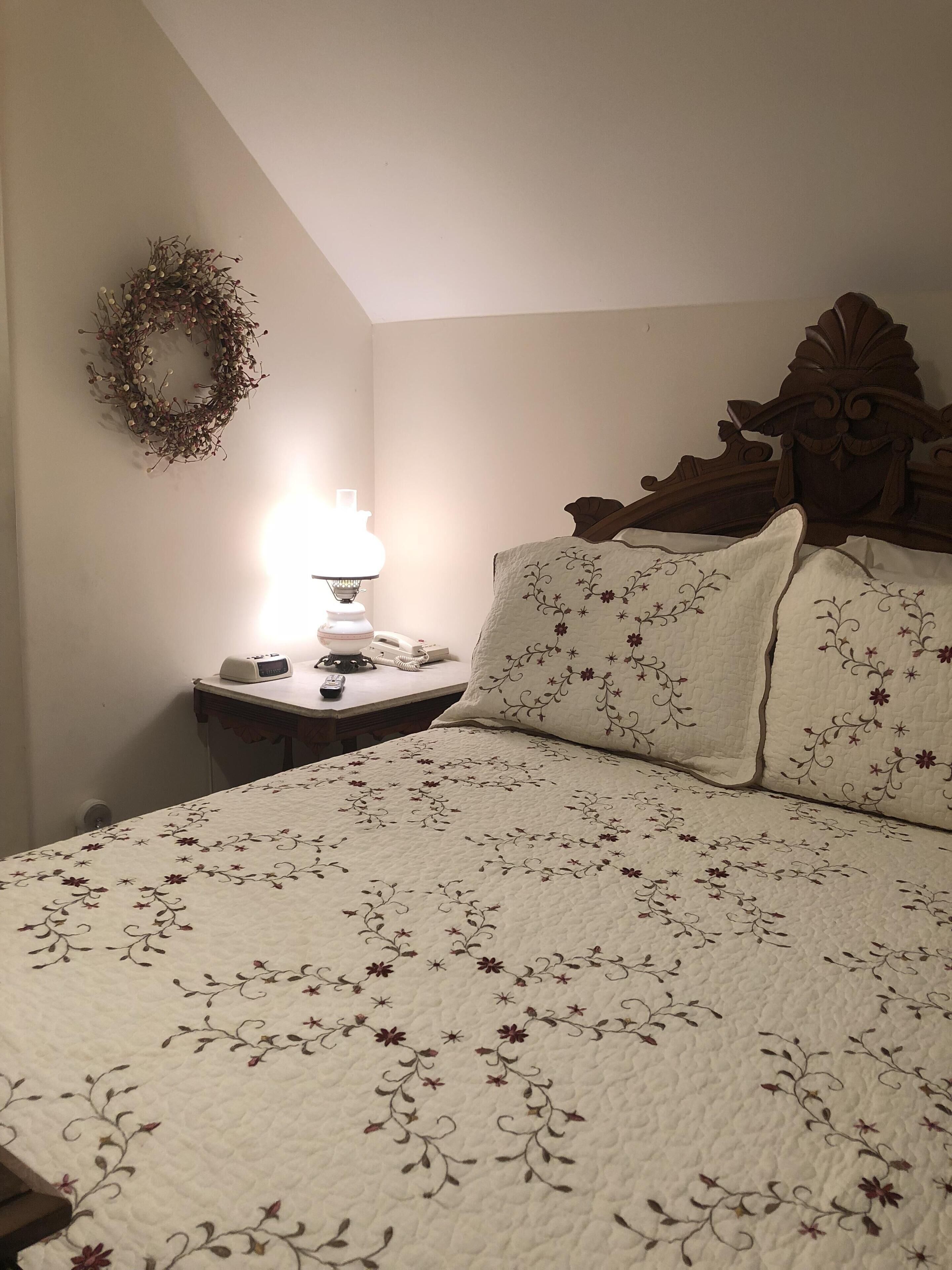 Un dormitorio con cama, mesita de noche, lámpara, teléfono y un arreglo decorativo en la pared.