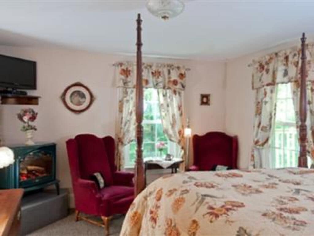 A bedroom with a four-poster bed, two red armchairs, a small table, and a television on a stand.