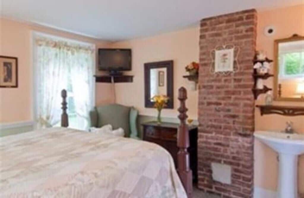 A hotel room with a bed, a brick accent wall, a television, a desk with a chair, and a window with curtains.