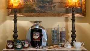 A table with a coffee maker, teapot, cups, and a decorative item against a wall with a framed painting. - Alpine Haus Bed & Breakfast Inn (Vernon)